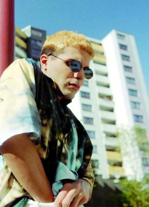 Ein Mann mit Sonnenbrille steht vor einem Hochhaus und greift in seine Hose.