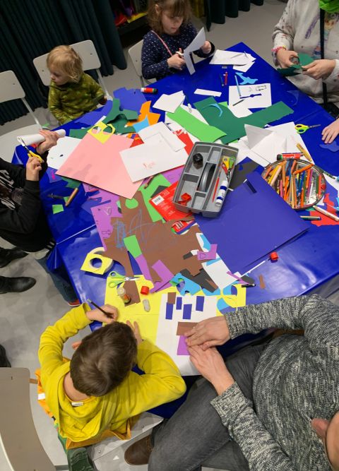 Man sieht viele Kinder an einem gro&szlig;en Basteltisch sitzen. Auf dem Tisch liegen viele bunte Bastelaccessoires.