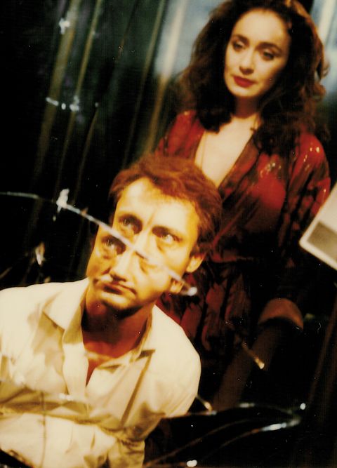 Ein Mann und eine Frau in einem Raum, der Mann sitzt vor einem zerbrochenen Spiegel und die Frau steht hinter ihm.