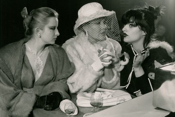 Drei elegant gekleidete Frauen sitzen auf einem Schwarzweißfoto nebeneinander, trinken Alkohol – eine spricht, zwei hören zu.