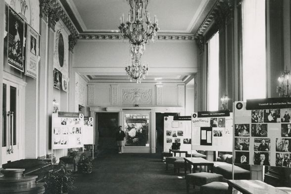 In einem historischen Raum mit Kronleuchter an der Decke stehen zahlreiche Stellwände mit Fotografien.