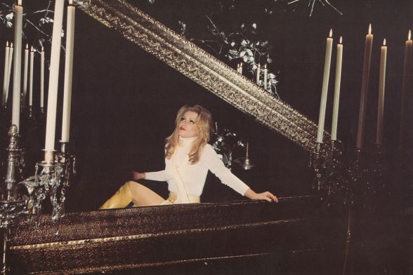 Eine blonde Frau sitzt in einem Pulloverkleid in einer Gruft, umgeben von Kerzenleuchtern.  A blonde woman sits in a sweater dress in a crypt, surrounded by candelabras.