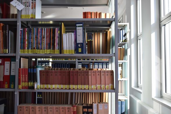 Bücher und Ordner stehen in einem Metallregal vor Fenstern.