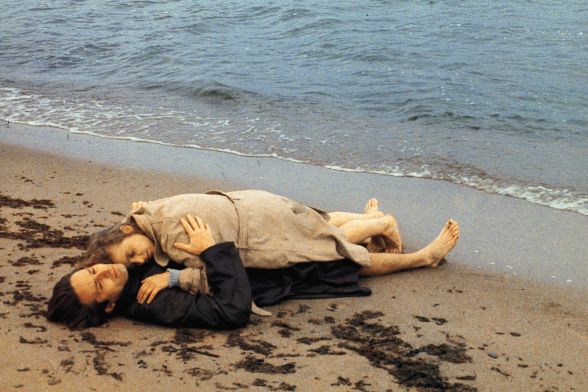Eine Frau liegt auf einem Mann am Strand, beide tragen Mäntel.