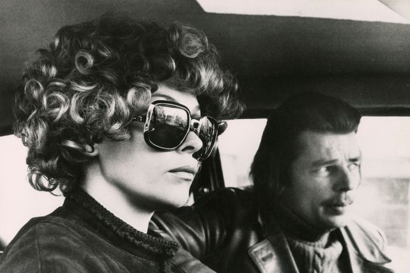 Eine Frau mit gro&szlig;er Sonnenbrille sitzt ernst neben einem Mann mit halblangem Haar und Schnurrbart im Auto.