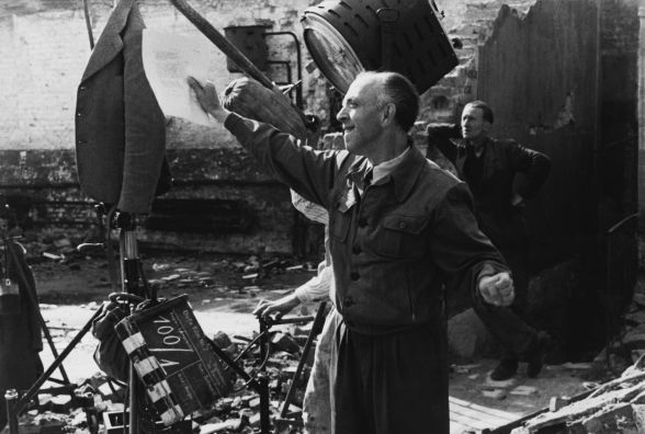 Der Regisseur steht in zerstörter Stadtkulisse und gestikuliert mit den Armen.