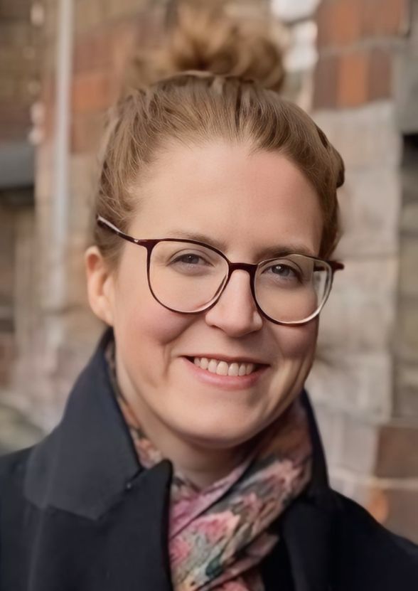 Eine Frau mit rotbraunem hochgestecktem Haar und Brille l&auml;chelt freundlich in die Kamera vor neutralem Hintergrund.