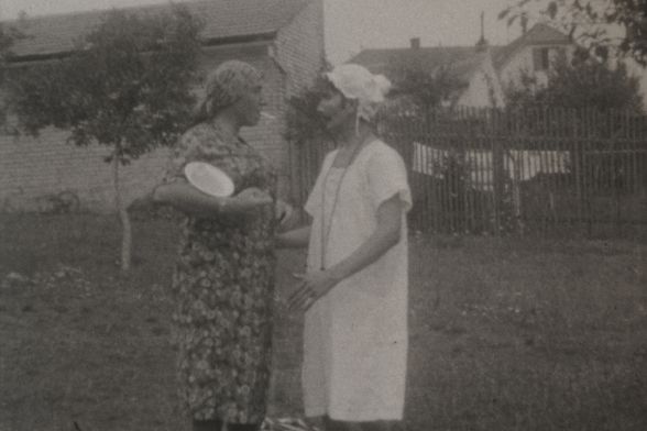 Two men in robes and caps are standing in a garden, appearing to be in a discussion.