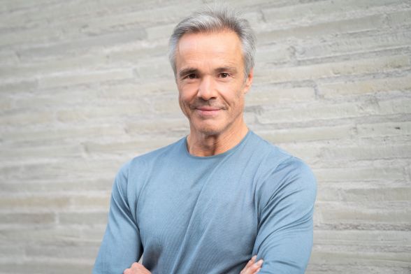 Der Schauspieler steht vor einem neutralen Hintergrund, schaut freundlich in die Kamera und hat die Arme verschränkt.