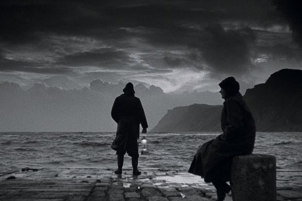 Eine Frau und ein Mann, beide in Regencapes, stehen am Meeresufer und blicken auf einen stürmischen Himmel voller Wolken.
