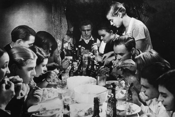 Mehrere Menschen unterschiedlichen Alters sitzen an einem Tisch, auf dem Bierflaschen, Schüsseln und Teller stehen, und essen.
