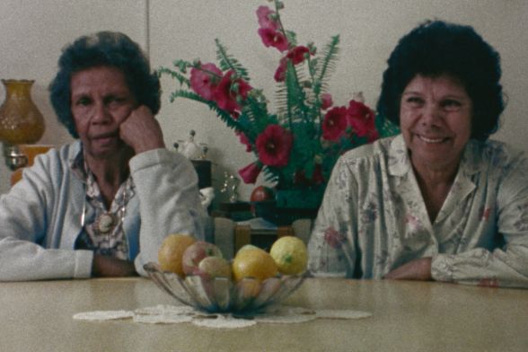 Two elderly women are sitting at a round dining table, one with a stern expression and the other laughing joyfully.