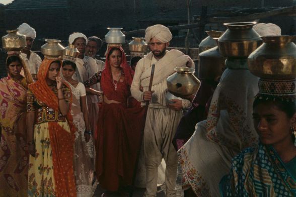People in saris and turbans, with vessels on their heads, look ahead in confusion.
