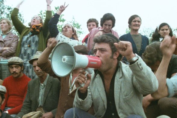 A man, surrounded by various people, is shouting something into a megaphone.