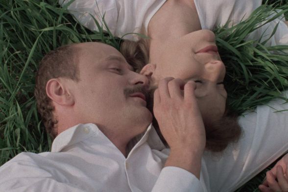 A man and a woman in white clothing lie side by side on a meadow, smiling at each other and touching.