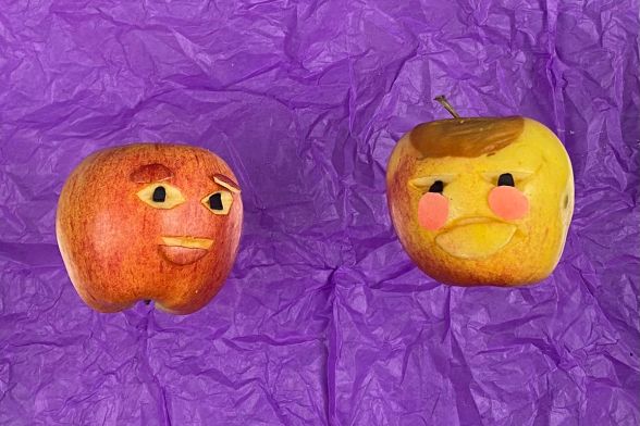 Zwei Äpfel auf lilafarbenem Papier wirken wie Gesichter: Augen und Mund sind eingeritzt, sie scheinen zu lächeln.