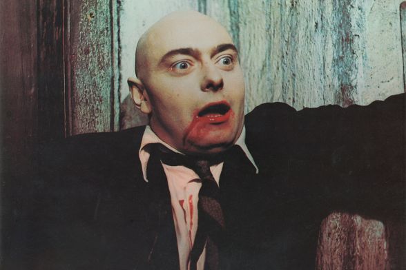 Ein Mann ohne Haare mit weit aufgerissenen Augen hat einen blutverschmierten Mund und Blut auf seinem weißen Hemd.