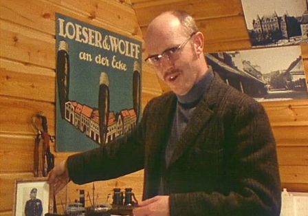 Ein Mann im Style der 60er Jahre, steht in einem Zimmer mit Plakaten an der Wand. Er spricht und zeigt ein Foto. 
