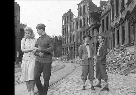Vier Personen in einer vom Krieg zerst&ouml;rten Stadt, links ein Paar, das die Stra&szlig;e entl&auml;ngl&auml;uft, rechts zwei M&auml;nner, die ihnen hinterherschauen.