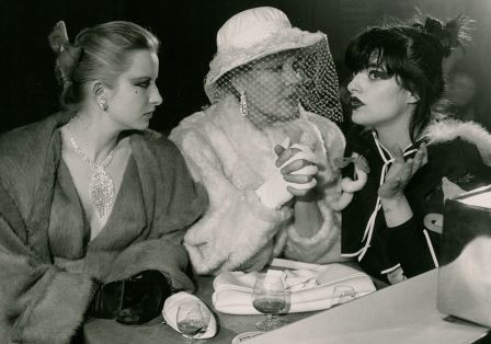 Drei elegant gekleidete Frauen sitzen auf einem Schwarzweißfoto nebeneinander und trinken Alkohol, eine spricht, zwei hören zu.