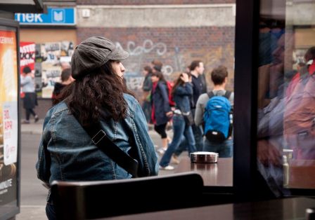 Eine Person mit langen Haaren sitzt auf einer Bank und schaut auf die Stra&szlig;e.