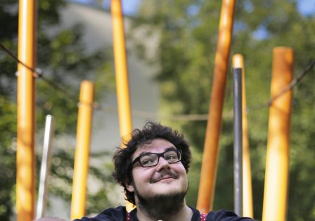 Mann mit Brille, der sich auf einen Tisch st&uuml;tzt und in die Ferne schaut, mit gro&szlig;en orangefarbenen Stangen im Hintergrund.