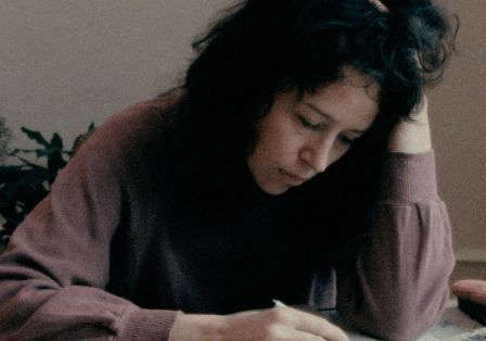 Eine Frau sitzt an einem Tisch und schreibt mit einem Stift. Sie hat ihren Kopf auf ihre Hand gest&uuml;tzt. 