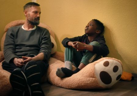 Ein Junge und ein Mann sitzen in einem Raum am Boden. Hinter dem Mann sieht man ein gro&szlig;es Teddyb&auml;r Kuscheltier. 
