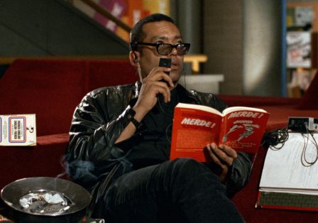 Ein schwarzer Mann mit Sonnenbrille sitz auf einer Couch. Er hat ein rotes Buch in der Hand und spricht auf ein Tonband. 