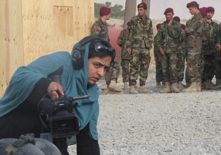 Eine Frau mit Kopftuch und Kamera befindet sich neben einer Menge von Soldaten. 