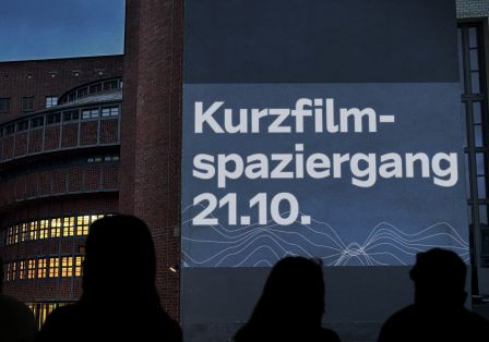 Menschen stehen draußen vor einer großen Leinwand mit dem Text „Kurzfilm-Spaziergang 21.10.“. Es dämmert und im Hintergrund ist das E-Werk mit beleuchteten Fenstern zu sehen. 