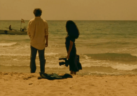 Ein Mann und eine Frau stehen am Strand und schauen auf das Meer, die Frau, rechts im Bild, h&auml;lt eine Filmkamera in der Hand.