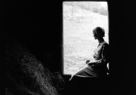 Eine Frau sitzt in einer Scheune am Fenster. Sie schaut nach drau&szlig;en und wirkt melancholisch. 