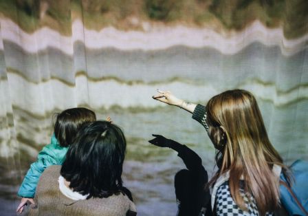 Eine Familie steht vor einem Vorhand auf den Streifenmuster projiziert sind. Ein Kind macht h&auml;lt seine Hand so, dass der Schatten einen Tierumriss auf den Vorhang wirft.