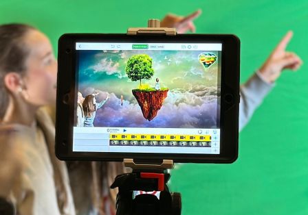 Kinder hinter einem Tablet auf einem Stativ, vor einem Greenscreen. Auf dem Tablet sind die Kinder in einer Landschaft zu sehen.