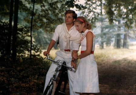 Zu sehen sind ein Mann auf dem Fahrrad und eine Frau an ihm gelehnt. Sie stehen im Wald und tragen wei&szlig;e Kleidung. 