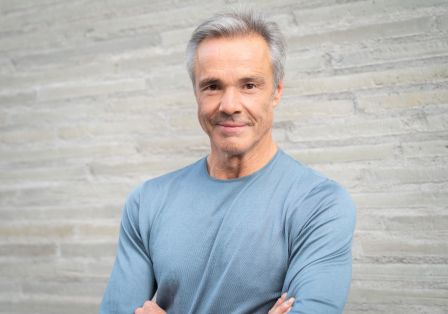 Der Schauspieler steht vor einem neutralen Hintergrund, schaut freundlich in die Kamera und hat die Arme verschränkt.