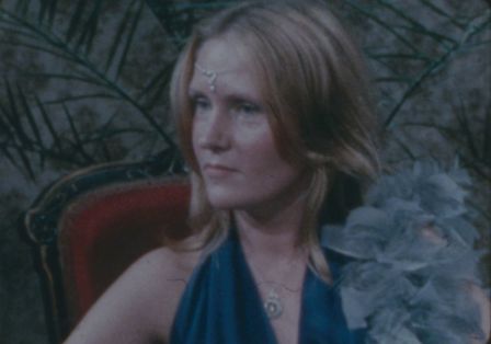 Frau mit blauen Kleid, sitzt auf einem Stuhl mit Bl&auml;ttern im Hintergrund.