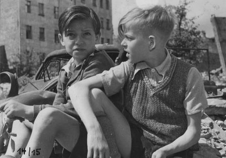 Zwei Jungen in kurzer Kleidung sitzen auf Tr&uuml;mmern in kriegszerst&ouml;rtem Berlin, mit Autowrack und zerst&ouml;rten H&auml;usern im Hintergrund.