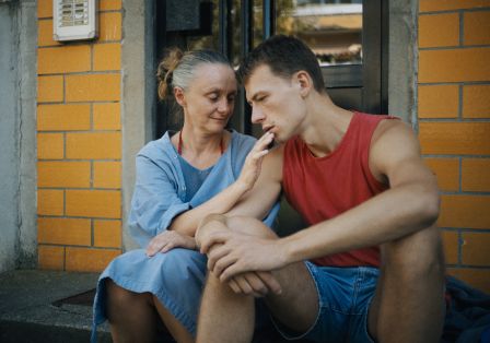 Man sieht einen jungen Mann und eine &auml;ltere Frau auf einem Treppenabsatz sitzen. Die Frau h&auml;lt ihre Hand an den Mund des Mannes. 