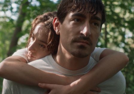 Eine junge Frau hängt schlapp auf den Schultern eines Mannes mit dunklen Haaren und Schnurrbart, der nach vorne schaut.