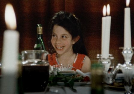 Ein kleines lachendes Mädchen sitzt zwischen Kerzenlicht an einem Tisch.