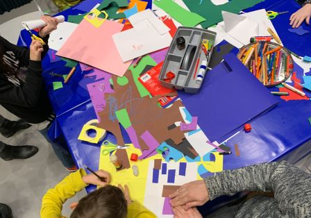 Man sieht viele Kinder an einem gro&szlig;en Basteltisch sitzen. Auf dem Tisch liegen viele bunte Bastelaccessoires.