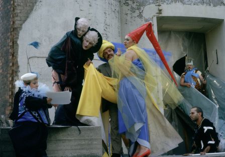 Mehrere Personen in bunten Kost&uuml;men stehen vor einem Geb&auml;ude und lesen etwas auf einem Blatt.