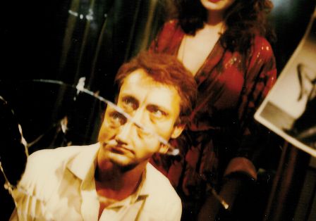 Ein Mann und eine Frau in einem Raum, der Mann sitzt vor einem zerbrochenen Spiegel und die Frau steht hinter ihm.