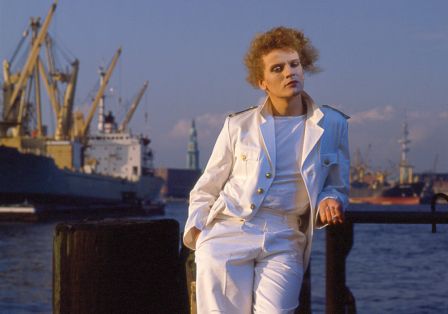 Person in wei&szlig;em Matrosenanzug steht am Wasser, im Hintergrund ein Hafen.