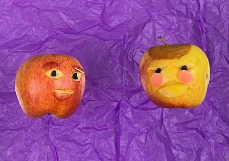 Zwei Äpfel auf lilafarbenem Papier wirken wie Gesichter: Augen und Mund sind eingeritzt, sie scheinen zu lächeln.