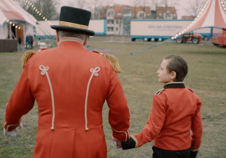 Ein Zirkusdirektor h&auml;lt einen Jungen in &auml;hnlichem Kost&uuml;m an der Hand und sie gehen gemeinsam in Richtung Zirkuszelt.