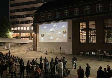 Es ist Abend und eine Menschengruppe steht vor zwei Häusern, ein Hochhaus im Hintergrund davor ein kleines älteres Haus auf dessen helle Außenwand ein Filmbild projiziert wird. 