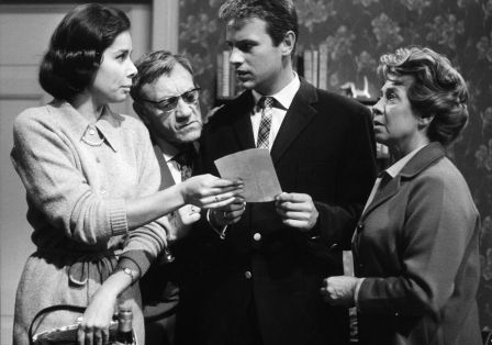 Vier Personen in den 1960er Jahren, die sich unterhalten und ein Blatt Papier betrachten.